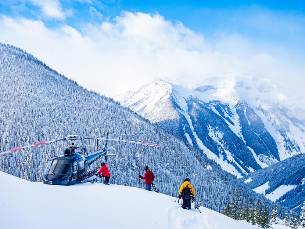 Skiiers approach a helicopter in front of snowy mountain peaks
