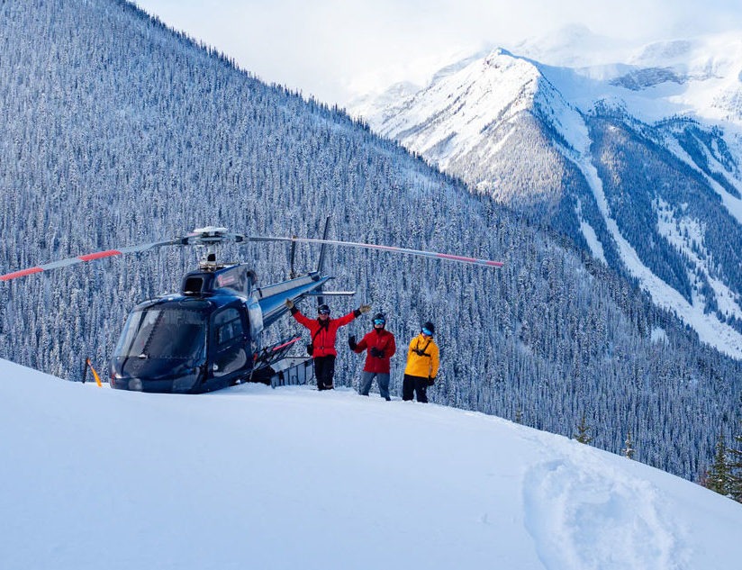 Skiiers wave next to a helicopter in front of snowy mountain peaks