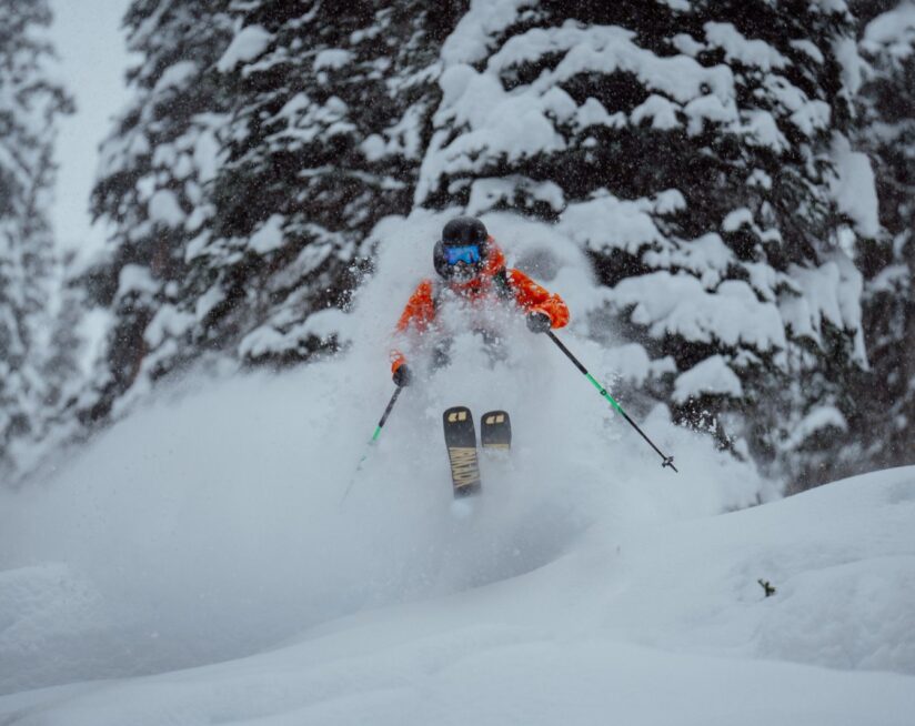 Skier charging through deep powder snow in a forested heliskiing zone