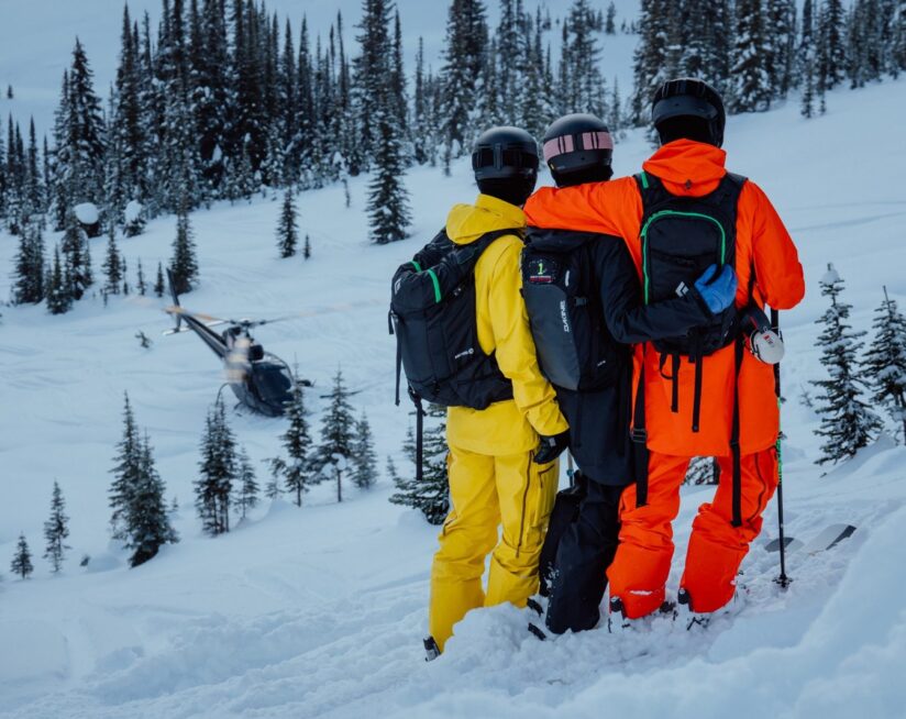 Three heliskiers overlooking a helicopter landing in a snowy alpine forest